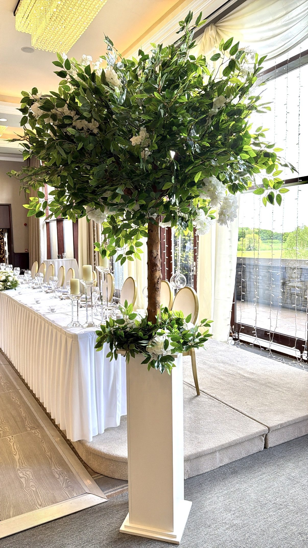 Luxurious green artificial tree on a white plinth beside a dressed top table